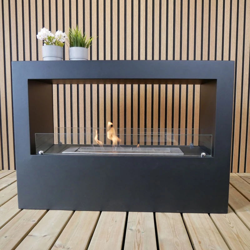 Sort firkantet bio pejs på træ terrasse med flammer i gang. Oven på bio pejsen står 2 planter i krukker.